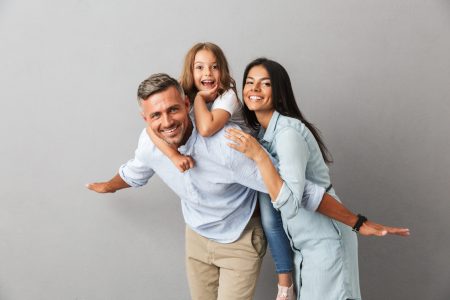 Portrait of a cheerful family father, mother, little daughter having fun together, father holding little daughter on his back isolated over gray background
